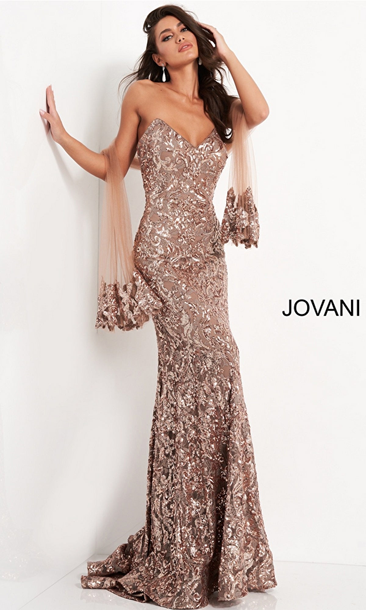 A woman poses in the Jovani Strapless Sequin Long Prom Dress 05054, a floor-length rose gold gown with a fitted silhouette and sheer tulle draping from her arms. The brand name
